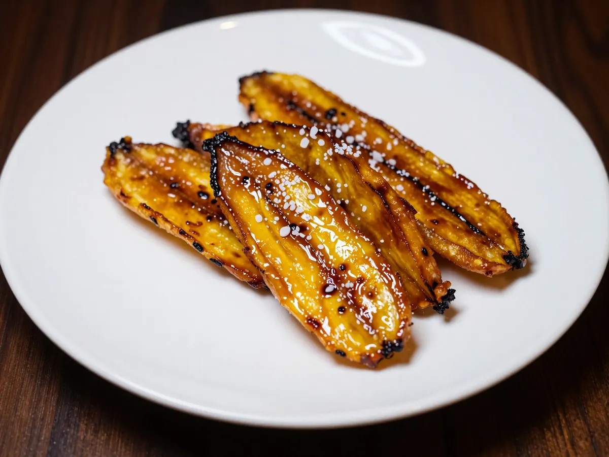 AI-generated photograph: Glossy slanted slices of caramelised fried plantain fanned in a neat overlapping arc on a matte white plate, edges deep amber, a few flakes of sea salt on top
